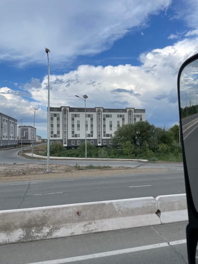 This is a photograph taken from a vehicle, likely in Qanlikul, Uzbekistan. The scene shows a modern residential building with white and dark gray facades, featuring numerous windows. The building is surrounded by lush green trees and vegetation. A paved road runs alongside the building, separated by a concrete barrier with white markings. Streetlights with solar panels are visible on tall poles. The sky is a vibrant blue with scattered white clouds, suggesting a clear and bright day. In the distance, more buildings and trees can be seen, indicating a developing urban or suburban area. The image appears to be taken during daylight hours. There is no visible text in the image.