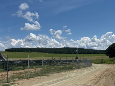 The image shows an outdoor scene with a clear blue sky filled with fluffy white clouds. In the foreground, a dirt road curves to the right, bordered by a metal chain-link fence. Beyond the fence, there are solar panels arranged in rows, set on a grassy field. A line of trees forms a dense forest in the middle ground, separating the solar farm from a larger green field beyond. A single large tree stands on the right side of the image, near the edge of the frame. A security camera is mounted on a pole to the left of the solar panels, suggesting this may be a secured area. The image appears to be taken in Verinsko, Bulgaria, a location known for its solar power generation.