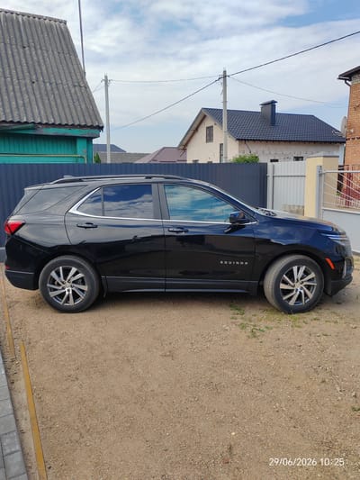 The image depicts a black Chevrolet Equinox parked on a gravel surface in Maladzyechna, Belarus. The car is positioned in a driveway, with a grey metal fence and a section of a building with a green painted wall on the left side of the frame. In the background, several houses with tiled roofs and fences are visible under a cloudy sky. A date and time stamp, "29/06/2026 10:25", is present in the bottom right corner of the image. The scene appears to be a residential area.