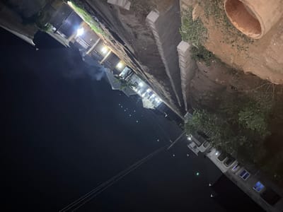 An off-kilter, nighttime view includes buildings lit with bright lights at the top. Below, a raised concrete structure parallels the buildings, transitioning into a dirt embankment. A cylindrical pipe rests on the ground above the embankment. The lower portion features more buildings with lit windows and a few trees. Power lines diagonally cross the dark foreground.
