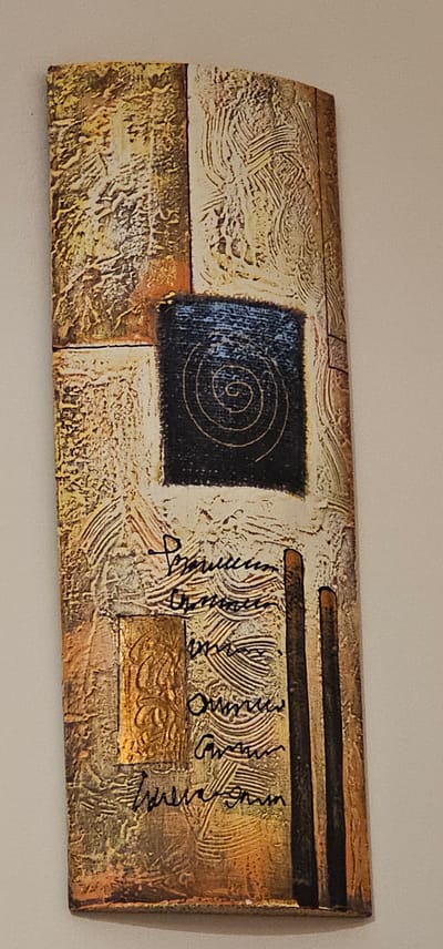 The image displays a close-up, vertical shot of a textured, abstract art piece mounted on a plain, light-colored wall. The artwork is elongated and appears to be a relief sculpture or a heavily textured painting. The palette is dominated by earthy tones: golds, browns, creams, and some black and rust orange.
The surface of the artwork is highly textured, with visible impasto and rough, almost geological formations of paint or plaster. There are distinct sections within the artwork. In the upper left, there's a block of textured golden-brown. To its right, a section of cream with parallel, raised lines that curve and intertwine, resembling wood grain or woven fibers.
A prominent feature is a square section of deep black in the upper-middle portion of the artwork. Within this black square, a light-colored spiral is inscribed, resembling a fingerprint or a galaxy. Below this black square, a large section of cream with similar curved, parallel lines is visible, interspersed with some reddish-brown accents.
Overlapping the textured cream sections, there is black, handwritten script. The script is illegible, appearing as abstract scribbles rather than clear text. It is strategically placed, adding another layer of visual interest and mystery to the piece.
Further down, a rectangular section of bright, textured gold is present. Below this gold rectangle, more of the cream-colored textured background is visible, along with further instances of the black, illegible script.
On the right side of the artwork, two vertical, dark brown or black rectangular elements are placed side-by-side, with a thin gap between them. These elements provide a stark contrast to the textured, lighter areas of the artwork.
The lighting in the image is soft and diffused, casting minimal shadows and highlighting the three-dimensional qualities of the artwork's surface. The wall appears to be a typical interior wall, likely in a home or gallery setting. There is no indication of the city of Dogato, Italy, within the image itself.