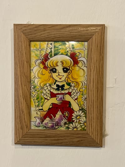 The image shows a framed illustration of an anime-style character, likely a girl, with blonde curly hair tied with red bows, large black eyes, and a red dress. She is holding a teacup and a teapot set is visible in the foreground, along with daisies. The illustration appears to be mounted on a wall, with a wooden frame. The wall is white and shows some minor imperfections and cracks. The lighting suggests an indoor setting. The provided context states this image is located in Maracay, Venezuela.