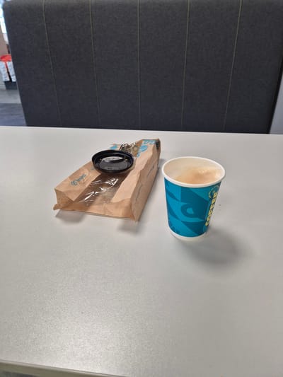 A close-up, eye-level shot shows a white table with a brown paper bag and a blue and white patterned coffee cup on it. The paper bag is partially open, revealing a clear plastic liner and a black lid resting on top. The coffee cup is full of coffee with a foamy top. In the background, a dark grey upholstered wall with vertical seams can be seen. The image is located in Ebavere küla, Estonia.