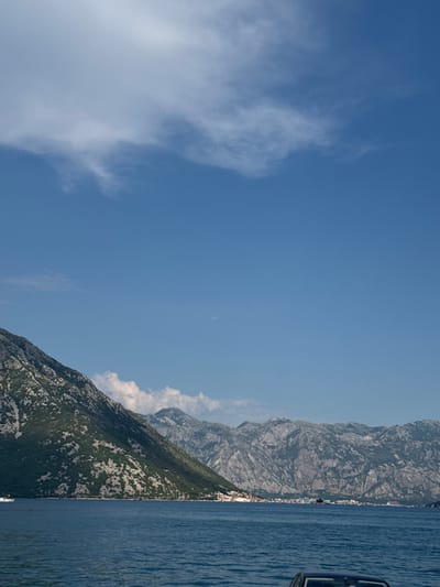 The image shows a body of water with a mountainous coastline under a blue sky with scattered clouds. A white boat is visible on the water in the distance on the left side. The mountains are covered with green vegetation and rocky outcrops. A town or village can be seen nestled along the shoreline in the middle distance. On the right side of the frame, a portion of a dark-colored car is visible in the foreground, partially submerged in the water, with its roof and windshield visible. The scene appears to be a scenic view from the water, likely in or near Kostanjica, Montenegro, given the characteristic bay and mountainous landscape.