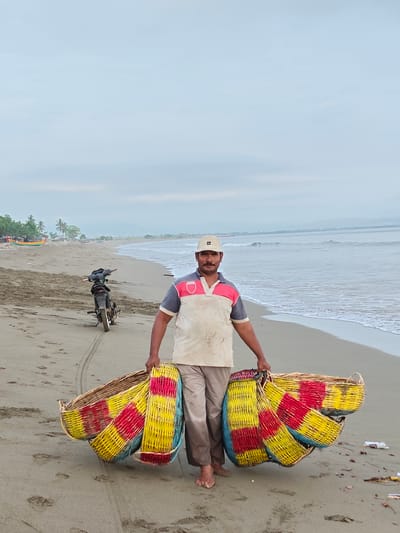 A man is walking on a beach in Curehtunong, Indonesia, carrying several woven baskets. He is wearing a light-colored polo shirt with red and grey accents and a light-colored baseball cap. His pants are a neutral tan color, and he is barefoot. He is holding multiple, colorful woven baskets, predominantly yellow and red, in front of him. Behind him, a motorcycle is parked on the sand. The ocean is visible to his right, with gentle waves rolling onto the shore. The beach is sandy with some debris scattered on it. In the distance, there are trees and a hazy mountainous landscape under a pale, overcast sky.
