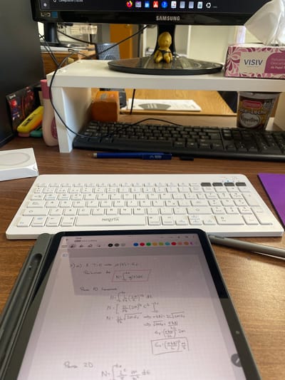 The scene depicts a desktop workspace located in San Carlos de Bariloche, Argentina. In the immediate foreground, a dark grey tablet is open, displaying handwritten mathematical equations and text in Spanish on a grid background, with "TP4 Teoria solidos" visible at the top. A grey stylus rests to the right of the tablet. Behind the tablet, a white NISUTA brand wireless keyboard is positioned, and further back, a black wired keyboard is present. A blue retractable pen, marked "NABA," lies between the two keyboards. To the left of the white keyboard, a small white box, possibly for an Apple MagSafe charger, is visible. The desk surface is light brown wood.

Centered in the midground, a black Samsung computer monitor is elevated on a white, two-tiered monitor stand. On the base of the monitor, a small golden-yellow figurine of a seated baby is placed. To the right of the monitor, a box of "VISIV" brand facial tissues and a lidded plastic container labeled "Bariloche Kapesche Dulce de Leche" are visible. To the left, a pink bottle and a multi-colored pen or tool are partially obscured. The monitor screen displays a desktop interface with multiple application icons and what appears to be a web browser. The overall environment suggests an office or study setting.
