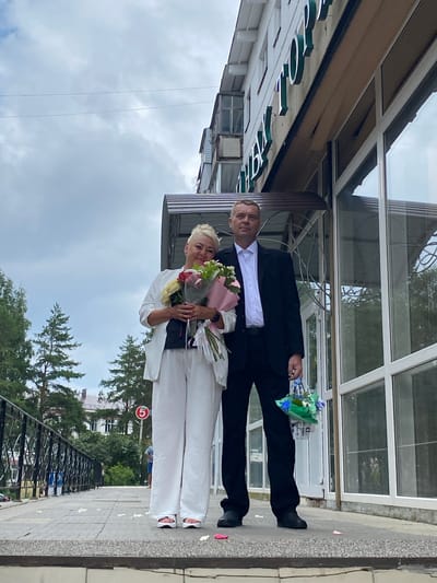 A man and woman stand on a sidewalk in front of a building. The woman is on the left, wearing a white jacket and white pants. She is holding a bouquet of flowers. The man is on the right, wearing a black suit and white shirt. He is holding a small basket of flowers. The building behind them has large windows and signage that appears to say "РЫБОЛОВ" which translates to "FISHERMAN" in English. A street sign with the number "5" inside a red circle is visible in the background. Trees and other buildings are visible further in the background. The sky is overcast. The location is the city of Chaikovsky, Russia.