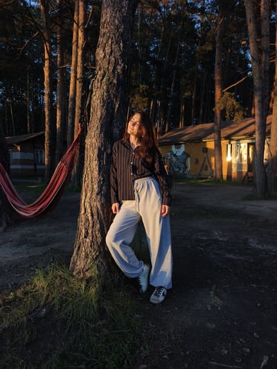 A young woman with long brown hair stands leaning against a thick tree trunk in a forest at sunset. She is wearing a striped long-sleeved shirt and light gray sweatpants. Her eyes are closed, and her face is tilted up towards the sun. To her left, a red and pink hammock is strung between two trees. In the background, there are two wooden buildings with roofs. The building on the left is a small, light-colored cabin with a dark roof. The building on the right is larger and yellow, with a mural of a man's face on its side. The sun casts long shadows through the trees, creating a warm, golden light. The ground is covered in pine needles and patches of grass. The location is the city of Онохино, Russia.