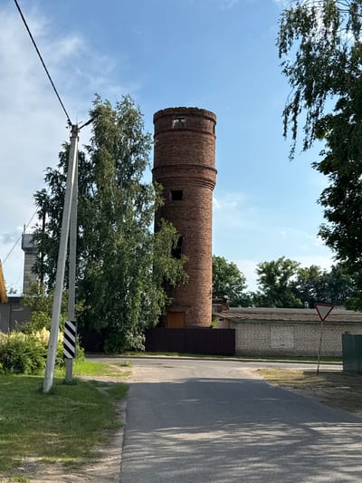 The image shows a red brick tower, possibly a water tower, with arched windows. The tower is surrounded by trees and greenery, with a road and a fence visible in the background. The sky is clear and blue, suggesting a sunny day. The overall scene appears to be in a rural or semi-rural area, possibly a village or small town. There are no people visible in the image. The photo is taken from a low angle, looking upwards towards the tower. The lighting and shadows suggest the time of day is mid-morning or early afternoon. The location is identified as Cherven, Belarus. There is a triangular warning sign visible on a pole, but no visible text.