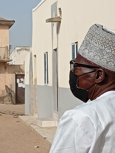 An individual is depicted in a right-profile view, positioned on a dirt path in Gayawa, Nigeria. The subject, a man with dark skin, wears a black face mask covering his mouth and nose, black-framed glasses, and a white and grey patterned cap. His attire includes a white long-sleeved garment featuring subtle striped and patterned textures. In the mid-ground, a light beige building with a grey lower section and two dark-framed windows is visible. A damaged concrete protrusion extends from its upper wall. Further back on the left, a light brown building with a sloped metal roof and a balcony with a metal railing is present. Adjacent to this, another off-white building with a dark wooden door covered in papers is discernible. The ground consists of uneven dirt and sand. The sky is clear blue.