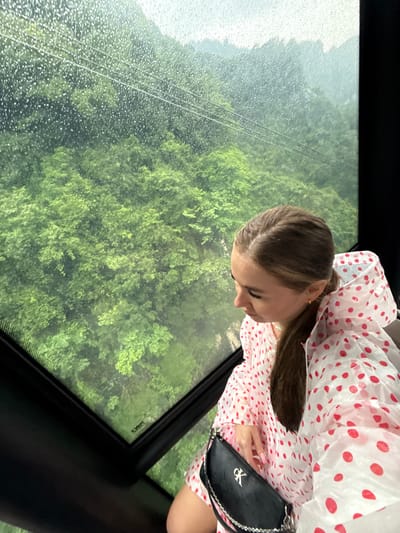 A woman is inside a cable car, possibly at Tianmen Mountain in Yongding District, China. Rain is visible on the cable car window, obscuring the view of the green, forested landscape outside. The woman has brown hair pulled back in a ponytail, and is wearing a white and red polka dot poncho. She has a small black purse with a silver chain.