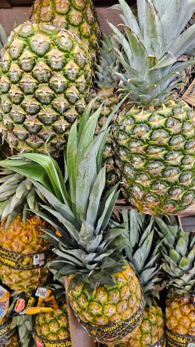 The image displays a close-up, overhead view of a display of Honeyglow pineapples at a market stall. The pineapples are stacked and arranged in a natural, abundant manner. Their characteristic spiky green leaves and textured, scaled exteriors are visible. Some pineapples have prominent green labels wrapped around them, indicating the brand and type. The lighting suggests an indoor environment, possibly a supermarket or farmer's market. There are no people or animals visible in the frame. The context provided places this scene in the city of Cuarte de Huerva, Spain, implying this is likely a grocery store or market within that location.