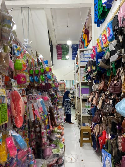 The image depicts the interior of a retail store in Lamteumen Barat, Indonesia, densely stocked with various merchandise. The store appears to have aisles lined with shelves and hanging displays. On the left side, racks are filled with packaged toys and kitchenware, including colorful plastic dishes and kitchen accessories. Further back, more shelves hold various items, and a woman wearing a hijab is seen examining products. The right side of the store features an extensive display of handbags and backpacks hanging from rods. These include a variety of styles, colors, and materials, such as leather, woven fabrics, and canvas. Shelving units behind the handbags contain boxes and additional merchandise. The lighting suggests an indoor environment, with overhead lights and a hanging fan visible. The floor is tiled.
