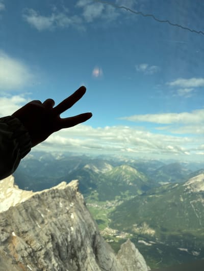An extreme close-up shot captures a silhouette of a hand making a peace sign against a vibrant blue sky dotted with fluffy white clouds. The hand, positioned in the lower left corner, is a dark shadow with its index and middle fingers extended upwards, forming the iconic "V" shape. The background transitions from the sky to a panoramic view of rugged, green mountains and a valley speckled with buildings, suggesting a scenic mountainous landscape. The image has a slight tilt, adding a dynamic feel to the composition. There are some minor smudges or reflections on the surface through which the photo was taken, particularly in the upper right quadrant of the sky. The overall impression is one of a person enjoying a breathtaking view from a high vantage point, possibly in Ehrwald, Austria, given the landscape's characteristics.