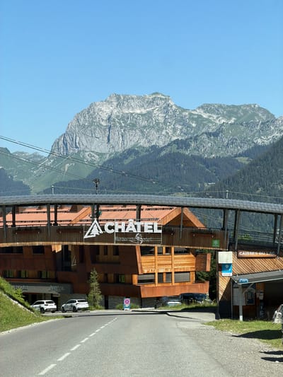 The image shows a road leading into the town of Châtel, France. On the left side of the road, there is a grassy embankment and a building with wooden siding and a large sign that reads "CHÂTEL". The sign is illuminated and has a stylized mountain logo. Further back, a covered walkway or bridge spans across the road, supported by wooden pillars. The building appears to be a resort or a hotel.

On the right side of the road, there is another building with a sign that says "CAISSES DU LAC". This suggests a ticket office or a point of sale, possibly for a lake or ski lift. Several cars are parked in front of the buildings, including two white SUVs on the left and a silver sedan and a black van on the right. In the background, a large, rugged mountain range dominates the skyline. The sky is clear and blue, indicating a sunny day. The overall impression is a scenic mountain resort town.