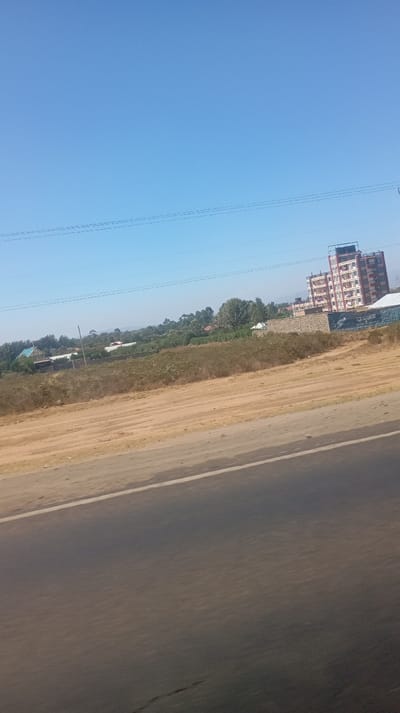 A roadside view with a highway occupying the lower portion of the frame. Beyond it, a dusty field transitions into a landscape dotted with shrubs and scattered houses. A mid-rise building with a red and white facade is prominent in the distance. An electric pole with wires stretches across the upper part. The sky is clear and bright blue.