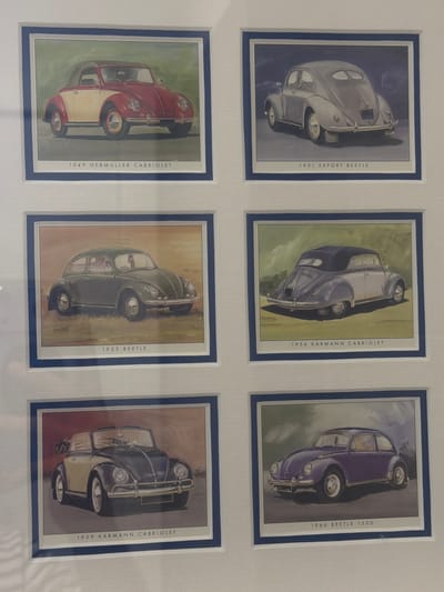 The image displays a framed collection of six framed prints, each featuring a watercolor painting of a Volkswagen Beetle. The prints are arranged in two columns and three rows. The top left print shows a two-tone red and white 1949 Hebmuller Cabriolet. To its right, the top right print depicts a silver 1951 Export Beetle. The middle left print showcases a dark green 1953 Beetle. The middle right print features a purple and white 1954 Karmann Cabriolet. In the bottom left, a yellow and dark blue 1959 Karmann Cabriolet is visible. Finally, the bottom right print displays a purple 1966 Beetle 1500. The framed prints are mounted on a white matte board, which is itself framed by a thin blue border. The overall scene is indoors, likely in a residential or gallery setting. There are no people or external environmental details such as city names or locations like Ouahat Sidi Brahim, Morocco, present in the image.