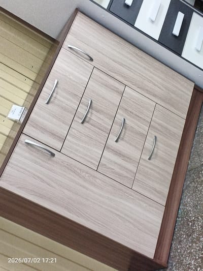 This media file shows a close-up, angled view of a piece of furniture, likely a cabinet or dresser, with a light wood-grain finish. The furniture has multiple drawers or cabinet doors, each with a sleek, silver handle. The doors are arranged in a grid-like pattern. To the left of the furniture, a price tag is visible, suggesting it might be displayed in a store. The background shows a beige, slatted surface and a section of a wall. The floor in the foreground appears to be concrete or a similar material with a speckled texture. The image is dated "2026/07/02 17:21," indicating it was taken in the late afternoon. There is no visible indication of the weather or any people. The overall scene depicts an indoor setting, possibly a furniture store or showroom in Chany, Russia.