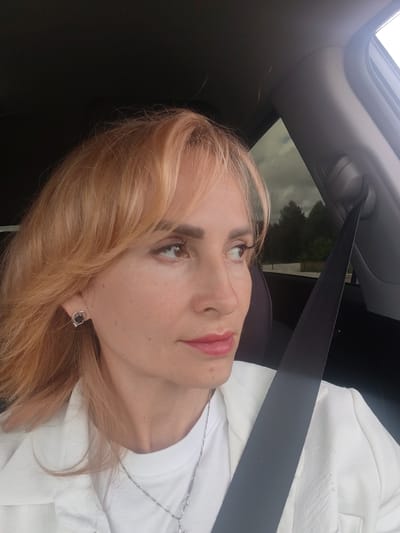 An unposed, close-up, medium shot shows a fair-skinned woman with blonde hair and fair skin sitting in the passenger seat of a car. She is wearing a white jacket over a white t-shirt and a silver necklace. A black seatbelt is draped across her body. She is looking out the window with a thoughtful expression, and her makeup is subtle. The interior of the car is visible in the background, as are trees and a cloudy sky through the window. The scene is likely Trofimovo, Russia, based on the provided context.