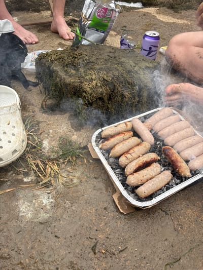 A disposable aluminum tray with sausages is positioned on a concrete surface near some moss and sparse grass. Some of the sausages are cooked more than others. There is a half-eaten package of sausages and a can next to the moss. Bare feet, a black sock, and a white Croc shoe are visible in the foreground. Smoke is rising from the moss.