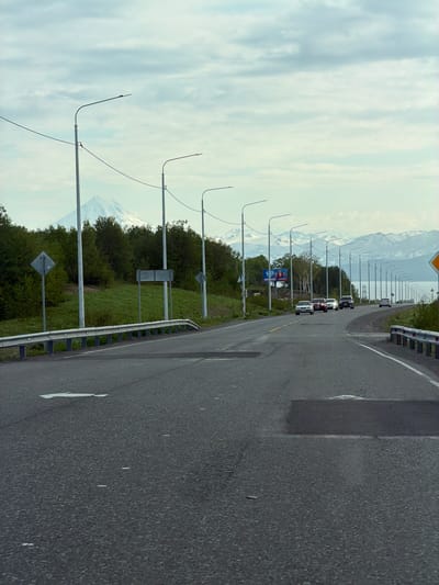 The image depicts a road in the city of Krutoberegovy, Russia, likely leading towards a mountainous or coastal area. The road is asphalted and appears to be a multi-lane highway with a guardrail on the right side. Several streetlights are visible along the side of the road, spaced at regular intervals. On the left side of the road, there is a grassy embankment with trees and a distant snow-capped mountain, possibly a volcano, in the background. A few cars are driving on the road, heading in the same direction. There are also some road signs, including a diamond-shaped sign on the left and a blank rectangular sign above it. The sky is cloudy with a hint of blue. The overall scene suggests a scenic route with natural elements and infrastructure.