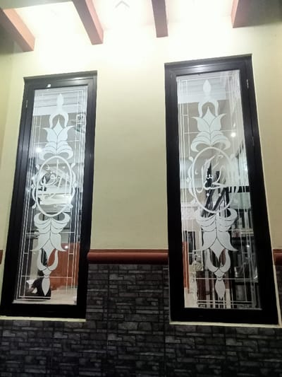 The image shows two windows with black frames and frosted glass. Both windows have an etched design in the center of the glass featuring a circular shape with Arabic script, surrounded by floral motifs. The window on the left has a person in a black shirt partially visible behind the glass. The windows are located on an interior wall of a building with a cream-colored painted surface. The wall is finished with a brown trim and a black brick veneer below. This location is in the neighborhood of Kecamatan Margorejo, Surabaya, Indonesia, with the coordinates -6.738 latitude and 111.014 longitude.