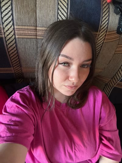 A close-up, top-down, portrait of a young woman is captured on a couch. She is lying on her side, facing the camera, with her head resting on a patterned sofa. She has medium-length, dark brown hair that falls around her face and shoulders. Her eyes are a striking green, and she is looking directly at the camera with a neutral expression. She is wearing a bright pink t-shirt, which is slightly creased. The sofa has a dark background with a woven pattern of thin horizontal lines in black, gold, and beige, with wider bands of blue and gold running vertically in sections. The lighting suggests it is daytime, and the scene appears to be indoors, likely in a home. There are no visible signs of text in the image. The overall mood is relaxed and casual.