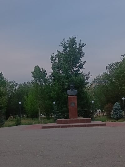 The image depicts a bust of a man in military uniform, wearing a cap, placed on a rectangular red stone pedestal. The pedestal is situated on a multi-tiered red brick platform, which is surrounded by a paved area in the foreground. Behind the monument, a dense arrangement of various trees, including tall, leafy deciduous trees and a conifer, forms a backdrop. Two white spherical lamps on poles are visible among the trees, suggesting a park or public space. The sky is overcast and a pale grey. The scene is devoid of any people or animals. The location is identified as Gvardeysky, Kazakhstan.