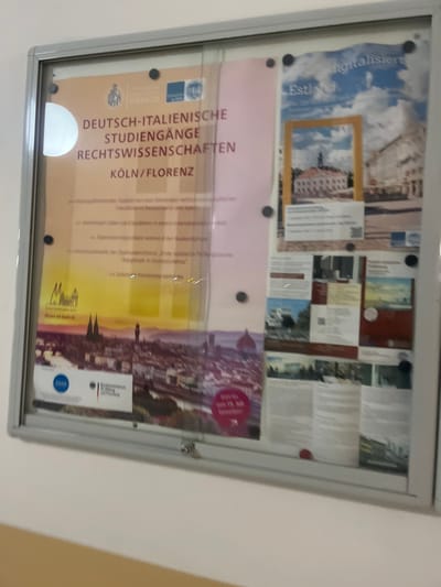The image shows a glass-framed bulletin board with several posters and flyers. It is located in Cologne, Germany, at the coordinates 50.929 latitude and 6.929 longitude. The bulletin board is mounted on a wall painted in a light beige color. The posters and flyers are all in German. The most prominent poster features the words "Deutsch-Italienische Studiengänge Rechtswissenschaften Köln/Florenz" and a blurred image of a city skyline. The poster features the logo of the University of Cologne and the logo of the University of Florence. There are several other posters and flyers, including one with a map of Europe and one with a photo of a building. The bulletin board is in a hallway. It is not possible to see what kind of building it is from the image, or any other people in the room. There are black dots on the glass that hold the posters and flyers. The bulletin board is angled slightly away from the camera. The bottom of the frame is mostly cut off by the bottom of the image. The image is blurry, but it is possible to make out the details of the posters and flyers.