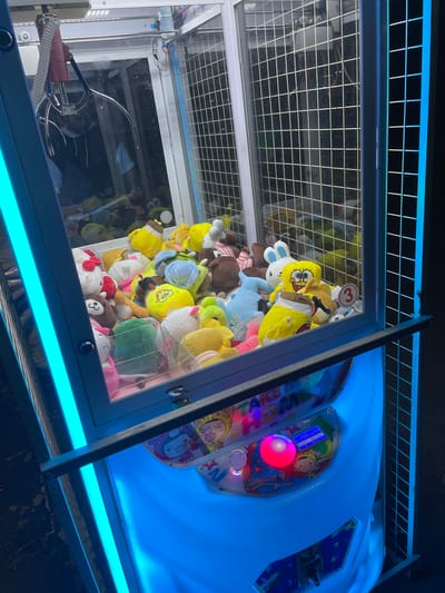 The image shows a claw machine filled with plush toys, predominantly yellow ones resembling Spongebob Squarepants, with other characters like Hello Kitty and a brown bear also visible. The machine is brightly lit with blue neon lights. There is a control panel with a red button and a digital display. The overall scene is indoors, likely in an entertainment venue. The city of Nova Zagora, Bulgaria, is not depicted or referenced in the image.