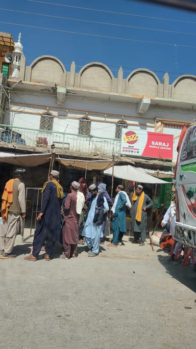 This daytime image captures a bustling street scene in Kuchlak, Pakistan. A group of men, dressed in traditional shalwar kameez and various types of head coverings such as turbans and caps, are gathered and walking along the side of a paved road. Their attire features a mix of colors including maroon, blue, green, and beige.

On the left, a building with distinctive arched architectural details along its roofline stands against a clear, bright blue sky. Multiple utility lines are visible crossing the sky. The building houses commercial establishments, indicated by visible signage. A prominent red sign reads "SARAJ MOBILE CENTER" in English. Below it, a yellow and red sign displays the "Jazz" logo along with the text "MERI SUPERPOWER" and several small circular icons. Part of a green and white vehicle, possibly a truck or bus, is parked near the shops. The street surface appears to be gray asphalt or concrete, brightly lit by the sun, suggesting a clear day.