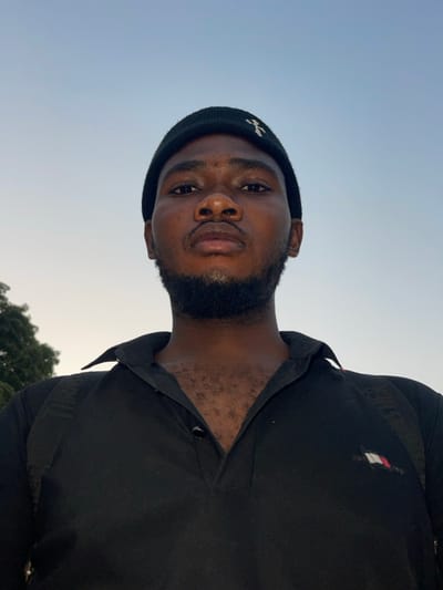 A forensic examination of the image reveals an adult male subject positioned outdoors in the city of Chikaji, Nigeria. The individual, presenting a dark complexion, gazes directly at the viewer with a neutral facial expression. Distinguishing features include a full, dark beard and mustache. He is attired in a black knitted beanie displaying a white cross or 'T'-shaped emblem on the front. Below, he wears a black collared polo shirt, with the top two buttons visible; the upper button is fastened, while the second is unfastened, partially revealing dark chest hair. A small, indistinct logo, resembling a rectangular flag, is present on the left chest of the shirt. Black straps consistent with a backpack are visible over both shoulders. The background consists primarily of a clear, light blue sky, with a blurry section of green foliage visible in the lower-left quadrant of the frame. The lighting is soft and even.