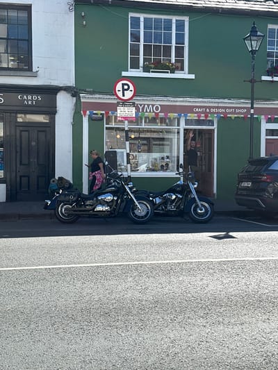 This is a vertically oriented, outdoor photograph taken in Westport, Ireland, on a sunny day. The image features two parked motorcycles, likely Harley-Davidsons, positioned on a paved street in front of a green building that houses a "CRAFT & DESIGN GIFT SHOP" called "Kymo". Above the shop entrance, a banner displays colorful triangular flags. To the left of the shop is a white building with a sign that reads "CARDS ART".
A person, appearing to be a woman with blonde hair, is leaning against the front motorcycle, holding a pink and white patterned item. Another person, a man with dark hair, is visible through the shop window, leaning out and gesturing with his hands.
A prominent "P" sign, indicating parking, is mounted on the green building, along with a "PAY & DISPLAY" notice. Visible text on signs includes "PHONE WATCH", "ONE HOUR FREE", and business hours. A dark-colored car is parked behind the motorcycles and in front of another building. The scene suggests a moment of pause or rest for the motorcyclists during their travels. The bright sunlight casts shadows, indicating it is likely daytime.