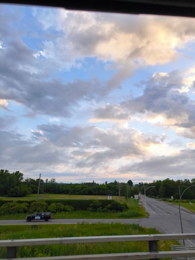 The image shows a highway scene in Saint-Lazare, Canada, with a black pickup truck driving on the road. The truck has "PUREY" written on its side, along with other decals. The road is a two-lane highway with grassy shoulders and trees lining the sides. The sky is cloudy with patches of blue and sunlight breaking through. In the background, there are more trees and some distant buildings. A railing is visible in the foreground, suggesting the photo was taken from an elevated position, possibly a bridge or overpass.