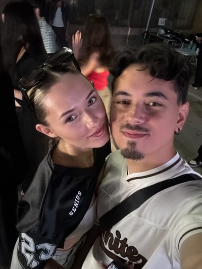 In Busmantsi, Bulgaria, a close-up selfie features a young woman with blonde hair pulled back, wearing sunglasses perched on her head and a black t-shirt with "GENIUS" written in white. She is looking towards the camera, her left side is partially obscured by shadows. Beside her, a young man with dark, curly hair and a goatee, wearing a white t-shirt with brown stripes and a black strap across his chest, smiles at the camera. They are in an outdoor setting with other people in the background, some of whom are out of focus. The overall lighting suggests it is nighttime or dimly lit.