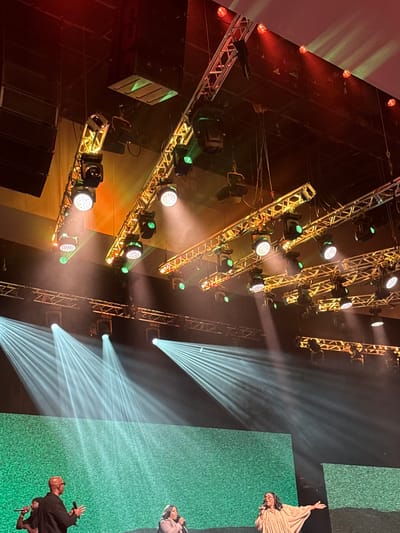The image depicts a stage setup, likely in Dallas, Texas, USA, with three individuals visible on stage. The backdrop is a textured green screen or wall. In the foreground, a man with a shaved head and glasses is speaking into a microphone, holding it with his right hand while gesturing with his left. To his left, another person is partially visible, also with a microphone. To the right, a woman with long dark hair and glasses is singing into a microphone, wearing a light-colored, flowing garment.
Above the stage, a complex lighting rig is suspended from trusses. Numerous spotlights and stage lights are visible, some emitting bright white beams that create dramatic rays in the smoky atmosphere, while others cast warm orange and red glows. Large black speaker arrays are also mounted above the stage. The overall impression is of a live performance or event in progress.