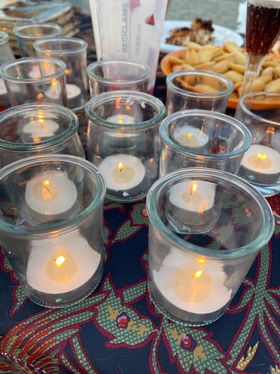 A close-up, overhead shot shows multiple lit tea candles in small glass jars, arranged on a dark blue tablecloth with a red, yellow, and green floral pattern. The candles glow warmly, casting light onto the surrounding glass and fabric. In the background, partially visible, are plates of food, including what appears to be fried potatoes and some kind of meat, as well as a glass filled with a dark beverage. A white container with text in what looks like French is also visible. The lighting suggests an outdoor evening setting. The scene is reminiscent of an al fresco dining experience.