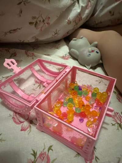 This image appears to be taken indoors, likely on a bed. The floral patterned bedding is predominantly white with pink and green floral designs. A pale-skinned person's leg and arm are present on the left side of the frame. Next to the leg is a small gray toy figurine, resembling a stylized animal with large eyes. To the right, a pink toy treasure chest is open. Inside the chest, there are a number of colorful water beads. The background is blurred, giving the image a shallow depth of field. The location is not discernable from the image.