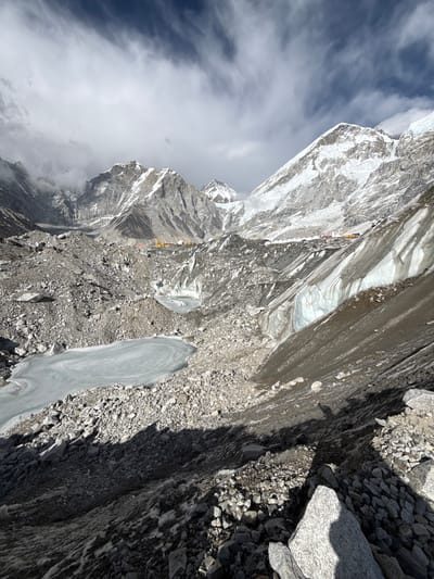 The image depicts a high-altitude mountain landscape, likely in the Khumbu region near Khumjung, Nepal, characterized by rugged terrain and glacial features. If viewed upright, the scene unfolds with a vast, rocky foreground transitioning into a challenging path through ice and rock formations.
In the mid-ground, several individuals, likely mountaineers or trekkers, are visible traversing a path. They are dressed in brightly colored outdoor gear, with visible colors including yellow, red, blue, and purple, standing out against the grey and white environment. These figures appear small, emphasizing the immense scale of the mountains. Further along the path, a cluster of yellow and orange shapes suggests the presence of tents, possibly indicating a high-altitude camp.
The landscape itself is a mix of dark, jagged rocks, scree, and patches of white and blue glacial ice. A distinctive, elongated, smooth ice formation is prominent in the foreground. The background features towering, snow-capped peaks partially enveloped by wispy white clouds, contrasting with a darker, clear blue sky above.
The image appears to have been taken during daytime, evidenced by clear shadows cast by rocks and terrain, suggesting direct sunlight. The weather seems partly cloudy but generally clear, allowing for expansive views of the mountainous expanse. The overall impression is one of challenging adventure and the formidable beauty of the Himalayan landscape. No text is visible in the image.