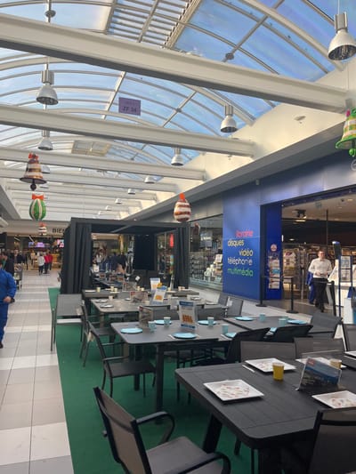 The image shows a view of an indoor shopping mall in Saint-Brice-Courcelles, France. The image shows a large, open-air shopping mall, featuring an open-plan design with a glass roof. There are several shops on either side of the mall, with a central walkway paved in black and white tiles. On one side of the walkway, a display of outdoor furniture is set up. This display consists of four black tables, six gray chairs, and a green rug. There are also several price tags attached to the tables. The space is decorated with festive Christmas decorations, including bells hanging from the ceiling. The mall is lit by multiple overhead lights. On the opposite side of the walkway, people can be seen walking, and several shops can be seen, one of which appears to be a multimedia store. A man is seen talking to a cashier. There is a sign in the store window that reads "Librairie, Disques, Video, Telephonie, Multimedia". A glass door leads into the store, revealing a well-lit and stocked interior. The store windows and doorways are guarded by black metal poles. There is a sign reading "Bien" in the distance.