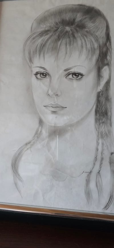 A black and white pencil sketch of a woman's face is framed in a thin silver and black frame. The drawing shows the woman from the shoulders up, with her head slightly turned to the left. She has dark, voluminous hair styled in a high updo with bangs. Her eyes are large and expressive, with visible eyelashes. She has a small nose and full lips. Two braids hang down on either side of her shoulders. The sketch is detailed, with shading used to create depth and dimension. The paper appears slightly creased and aged. The background is plain, with no discernible details. The frame rests on a dark wooden surface, possibly a table or shelf. There is no indication of a specific location in Bad Marienberg, Germany, within the image itself.