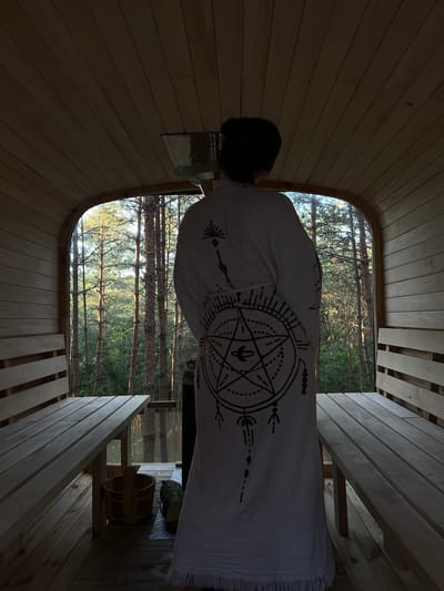 A person stands inside a wooden sauna, facing away from the camera and towards a large arched window that looks out onto a dense forest. The person is wearing a white robe with a large, intricate black symbol on the back. The sauna is constructed of light-colored wood, with benches on either side and a wooden floor. A wooden bucket and a log are visible on the floor. The natural light from the window illuminates the trees outside, creating a serene and natural atmosphere. The location is identified as Ivachiv Horishnii, Ukraine.