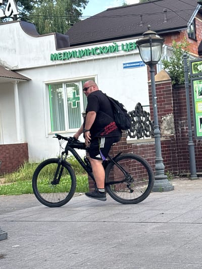 A bald man wearing sunglasses, a black t-shirt, black shorts with purple and white accents, and black athletic shoes is riding a black mountain bike on a paved sidewalk in Il’inskiy, Russia. The man has a black backpack on. The bike has a black frame, black wheels, and black tires. There is a black bag attached to the frame of the bicycle. The man is wearing a watch on his left wrist. He is looking to his right.

Behind the man, there is a white building with green signage that reads "МЕДИЦИНСКИЙ ЦЕНТР" (Medical Center). To the right of the building, a street sign with the text "ул. Московская 27" (Moskovskaya Street 27) is visible, indicating the location as Il’inskiy, Russia. A vintage-style street lamp with a dark metal base and a glass lantern is situated next to the street sign and in front of a red brick wall. The wall has an ornate black metal fence section. Green grass and some small white flowers are growing along the edge of the sidewalk and the brick wall. The sky is partially cloudy.