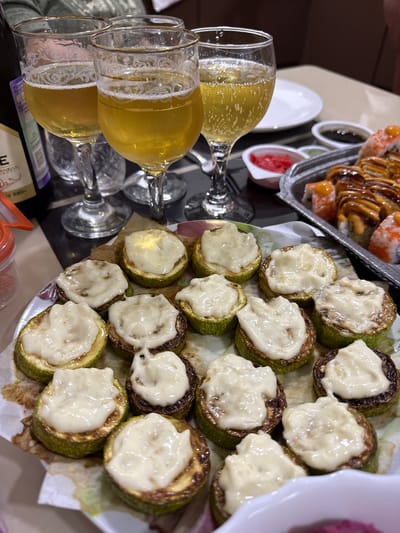 Three glasses of light-colored beer or sparkling wine are visible in the foreground, with a bottle partially obscuring the left glass. A platter of sliced zucchini topped with melted cheese is in the center, and a tray of sushi rolls is to the right. Small dishes of pickled ginger and soy sauce are also present. The scene appears to be a meal or celebration taking place indoors, likely in a home or restaurant setting. No specific location within Ishimbay, Russia can be determined from the image.
