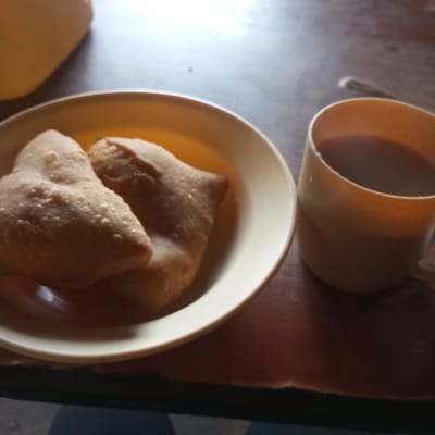 The image depicts a table in what is likely a home in Sheywe ward, Kenya. A beige plate contains two fried dough pastries. Adjacent to the plate is a tan-colored mug filled with a dark liquid, presumably coffee or tea. The table appears to be made of dark wood. Part of a yellow container is visible in the upper left corner of the frame. A blurry section of blue clothing is visible at the bottom of the image.