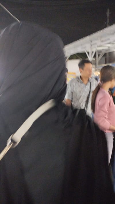 The image captures an outdoor scene at night, likely in Sukodono, Indonesia, based on the provided context. In the foreground, a person is seen from behind, wearing a black garment, possibly a hijab or a dark jacket, with a white strap across their shoulder. The background is somewhat blurred, typical of low-light photography or a moving subject.
Further back, two other individuals are visible. A man in a patterned, light-colored short-sleeved shirt stands in the middle ground. To his right, a woman with long, dark hair is wearing a pink long-sleeved top and grey pants. They appear to be walking or standing in a public space, possibly a market or a street fair, given the faint lights and structures visible in the background. A white awning or roof structure is prominent above them, suggesting an area with temporary stalls or shelters. The overall atmosphere is casual, and the dim lighting indicates it is nighttime. No text is discernible in the image.