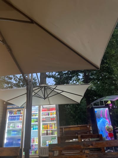 An outdoor scene in Nova Zagora, Bulgaria, under a large tan umbrella, features two illuminated beverage refrigerators filled with various bottled drinks. To the right, a large screen displays a blue animated character, next to a gumball machine. Wooden benches are visible in the foreground, with trees and foliage forming the background.