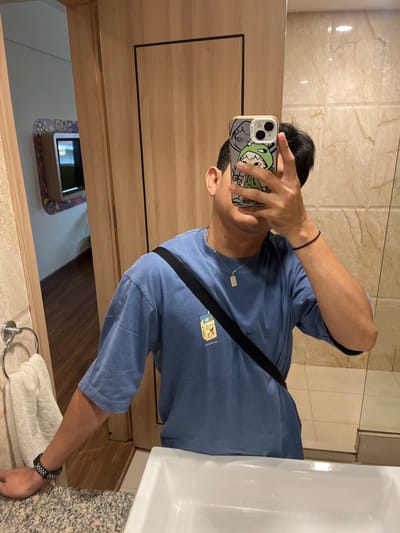 A person is taking a mirror selfie in a bathroom. The person is wearing a blue t-shirt with a graphic print and a black crossbody bag. They are also wearing a silver pendant necklace and a black bracelet on their right wrist and a watch on their left wrist. The bathroom has beige marble walls, wooden doors, and a white sink. A towel is hanging on a rack on the left side of the image. In the background, a small television is visible.