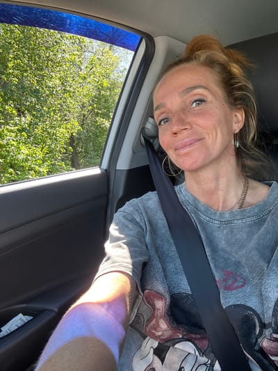 A woman is seated in the driver's side of a vehicle in Beloostrov, Russia, taking a selfie. Her light-colored hair is pulled back into a bun. She has blue eyes, a light complexion with visible freckles, and is smiling faintly towards the camera. She wears a faded gray t-shirt with a partial graphic design, a multi-strand gold-colored chain necklace, and gold-colored hoop earrings. A black seatbelt is positioned diagonally across her chest and shoulder. Her left arm, resting on the car's door panel, is strongly illuminated by direct sunlight, displaying a visible tan line and a purple/orange discoloration on the skin. The interior car door panel is dark gray or black plastic, with a small storage compartment holding a white paper slip, possibly a receipt. Outside the car window, dense green foliage from trees is visible under bright sunlight, with a small patch of blue sky visible at the top right. A blue wind deflector or tint is affixed to the upper edge of the car window.
