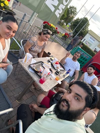A group of people are gathered around a table outside a McDonald's restaurant in Mainburg, Germany. A woman in the foreground on the left, wearing a white top and blue jeans, is holding a smartphone. To her right, a woman in a patterned dress is seated. In the center of the image, a man in a white t-shirt and shorts is holding a cigarette, looking towards the camera. A young man in a white tank top is seated to his right. The table is cluttered with McDonald's food containers, cups, and napkins, suggesting they have recently finished a meal. In the background, other individuals are visible, along with the McDonald's building and signage, as well as some trees and a playground structure. The overall scene depicts a casual outdoor dining experience.